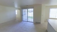 This single-floor residential space offers a straightforward and functional layout designed for comfortable everyday living. The main living area is bright and open, featuring wall-to-wall carpeting that adds warmth underfoot. A large sliding glass door with vertical blinds serves as a focal point of the living room, flooding the space with natural light and providing a direct view of the surrounding greenery outdoors. A floor vent ensures efficient climate control throughout the room. An additional window on an adjacent wall further enhances the sense of openness and brings in supplemental natural light. The interior walls are finished in a neutral, light tone, creating a clean and versatile backdrop suitable for a variety of décor styles. A connected area is visible through an open passageway, suggesting a well-integrated floor plan that allows for easy flow between living spaces. The overall ambiance is calm and neutral, offering a move-in-ready canvas for personalization. The single-floor layout ensures accessibility and convenience, with all spaces situated on one level for seamless navigation throughout the home.