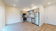 This two-story space features a clean, contemporary design with a neutral color palette and consistent modern finishes throughout. The main level is anchored by an open-concept kitchen and living area, where light wood-look luxury vinyl plank flooring runs seamlessly underfoot. The kitchen is well-appointed with light wood cabinetry, granite-style countertops, and a full suite of stainless steel appliances, including a refrigerator, electric range with microwave above, and dishwasher. Recessed lighting illuminates the space efficiently. Large windows fitted with horizontal blinds allow natural light into the living area, which offers an open, airy feel. A staircase leads to the upper level, where vaulted ceilings add architectural interest and a sense of vertical space. The upper-floor rooms are finished with plush carpeting, creating a comfortable and quiet atmosphere. The bathrooms, found on both levels, feature wood-look vinyl flooring, granite-topped vanities with wood cabinetry, and full tub-shower combinations with clean white surrounds and chrome fixtures. A hallway with multiple doorways connects key areas of the main floor, providing functional flow throughout the space.