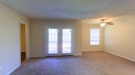 This single-floor residence offers a welcoming and functional layout designed for comfortable everyday living. The main living area is generously sized, featuring plush wall-to-wall carpeting that adds warmth underfoot. A standout feature of this space is the set of white French doors, which flood the room with natural light and provide access to an outdoor area, creating a seamless connection between interior and exterior. Blinds on all windows offer privacy while still allowing light to filter through. A ceiling fan is centrally positioned in the adjoining area, ensuring year-round comfort and air circulation. The walls are finished in a soft, neutral tone that complements the light-colored trim and baseboards, contributing to a clean and airy feel throughout. The textured ceiling adds a subtle architectural detail consistent across the space. The overall ambiance is bright, open, and practical, making it a versatile space well-suited for a variety of furnishing arrangements and lifestyles.