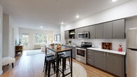 This thoughtfully designed 2-bedroom, 2-bathroom residence offers a modern, open-concept layout that seamlessly connects the kitchen, dining, and living areas. The kitchen is a standout feature, showcasing sleek gray cabinetry, white subway tile backsplash, stainless steel appliances including a range, microwave, and refrigerator, and light countertops. A high-top dining table with industrial-style stools provides a casual space for dining and entertaining. The living room is bright and welcoming, with large windows and a balcony door that invite natural light and offer views of lush greenery. Light-toned hardwood flooring flows throughout the main living areas, adding warmth to the contemporary design. The primary bedroom is a comfortable retreat, featuring plush carpeting, a closet with built-in shelving and hanging rods, and a window overlooking tree-lined surroundings. The second bedroom similarly offers carpeted flooring, natural light, and a dedicated closet. Both bathrooms are finished with wood-look flooring, white tub-shower combinations, and vanities with white countertops and gray cabinetry. A spacious walk-in closet with wire shelving provides ample additional storage. Recessed lighting throughout enhances the clean, modern aesthetic of this well-appointed residence.