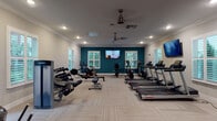 This fitness center offers a comprehensive workout environment designed to accommodate a wide range of exercise needs. The space is thoughtfully laid out across a single floor, with distinct zones dedicated to cardiovascular training and strength work. A row of Life Fitness treadmills lines one side of the room, accompanied by upright and recumbent bikes, cycling bikes, a rowing machine, and an elliptical trainer — providing ample options for cardio-focused workouts. On the strength side, a dumbbell rack stocked with a full range of hex weights, a weight bench, and a functional training rig offer versatile resistance training. A dedicated air purifier unit keeps the environment fresh throughout.

Architecturally, the room features elegant crown molding, recessed lighting, and dual ceiling fans that maintain comfortable airflow. A bold teal accent wall anchors the far end of the space, adding a vibrant, energizing visual element. Multiple windows fitted with plantation shutters allow natural light to filter in while offering views of surrounding greenery. Wall-mounted flat-screen televisions are positioned throughout for entertainment during workouts. The flooring consists of neutral, striped carpet tiles that lend a clean, modern aesthetic. A glass-panel exterior door provides convenient direct access to the outdoors, completing this well-appointed and functional fitness facility.