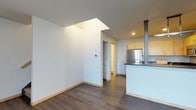 This three-story space offers a compelling contrast between refined modern finishes and raw industrial character. On one level, a clean, contemporary kitchen is equipped with light wood cabinetry, dark countertops, a stainless steel refrigerator, and a peninsula with a sleek range hood. Smooth white walls, wood-look flooring, and a skylight overhead introduce natural light and an airy, open feel. A built-in shelving nook and additional doorways suggest thoughtful storage and functional flow throughout.

The industrial level presents a dramatically different aesthetic, showcasing exposed brick walls, an open steel-and-timber ceiling structure with visible ductwork, and pendant lighting that hangs from considerable heights. Large industrial-style windows allow natural light to filter in, while a mezzanine or loft-style overlook with metal railing adds a sense of vertical depth. Polished wood bar-height tables and curved wood-and-metal stools anchor this communal area, lending warmth against the raw architectural backdrop.

Together, the floors create a layered, textured environment that balances contemporary comfort with industrial heritage, making it a versatile and visually engaging space.