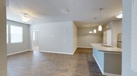 This single-story space showcases an open-concept layout that effortlessly connects the kitchen and living areas, creating a cohesive and functional environment. Rich, wood-look plank flooring runs throughout, adding warmth and visual continuity across the space.

The kitchen area features a teal-accented peninsula with a light stone countertop, offering both prep space and a natural gathering point. A stainless steel sink is integrated into the peninsula, and a set of stylish pendant lights — including a frosted glass fixture and a matching pendant — hang above, providing focused task lighting with an aesthetic touch. White cabinetry is visible along the perimeter.

The adjoining living area is generously sized, with neutral gray-toned walls and clean white baseboards contributing to a bright, modern feel. A ceiling fan is installed overhead, enhancing comfort. A textured ceiling adds subtle dimension throughout. Natural light filters in through a window visible in an adjacent room, which appears to connect seamlessly to the main space via an open doorway, further enhancing the sense of flow and openness throughout this single-level layout.