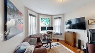 Located on Commonwealth Avenue in Boston's Allston neighborhood, this one-bedroom, one-bathroom unit offers a practical and characterful layout across a single floor. The living room is a standout space, anchored by a classic bay window that fills the room with natural light and features white-trimmed molding that adds architectural charm. Warm hardwood floors run throughout the unit, lending a cohesive and inviting feel to every room. The living area provides enough space to accommodate both a seating area and a dedicated workspace. The bedroom is a well-proportioned retreat highlighted by an exposed brick accent wall, two windows with views of the surrounding tree line, and the same rich hardwood flooring. Crown molding adds a traditional finishing touch. The galley-style kitchen is efficiently designed with wood cabinetry, a full-size refrigerator, and a sink with counter space on both sides, connecting directly to the rest of the unit via a short hallway. The bathroom is clean and bright, featuring white subway tile surrounding a tub-shower combination, a pedestal-style sink, and a window that allows natural light to filter in. A hallway with coat storage ties all rooms together, making the layout feel organized and functional throughout.
