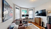 Located on Commonwealth Avenue in Boston's Allston neighborhood, this one-bedroom, one-bathroom unit offers a practical and characterful layout across a single floor. The living room is a standout space, anchored by a classic bay window that fills the room with natural light and features white-trimmed molding that adds architectural charm. Warm hardwood floors run throughout the unit, lending a cohesive and inviting feel to every room. The living area provides enough space to accommodate both a seating area and a dedicated workspace. The bedroom is a well-proportioned retreat highlighted by an exposed brick accent wall, two windows with views of the surrounding tree line, and the same rich hardwood flooring. Crown molding adds a traditional finishing touch. The galley-style kitchen is efficiently designed with wood cabinetry, a full-size refrigerator, and a sink with counter space on both sides, connecting directly to the rest of the unit via a short hallway. The bathroom is clean and bright, featuring white subway tile surrounding a tub-shower combination, a pedestal-style sink, and a window that allows natural light to filter in. A hallway with coat storage ties all rooms together, making the layout feel organized and functional throughout.