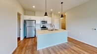 Located on S Burnsfield Ave in Sioux Falls, South Dakota, this single-floor unit offers a well-designed layout with modern finishes throughout. The kitchen serves as a central highlight, featuring a large island with a light quartz countertop and integrated sink, crisp white shaker cabinetry, stainless steel appliances including a refrigerator, electric range, and built-in microwave, and stylish pendant lighting — all set over warm hardwood-style flooring. The living and main areas continue the hardwood-style flooring and are filled with natural light from generously sized windows offering open views of the surrounding landscape. Exposed ductwork adds an industrial design element to the space. Bedrooms are carpeted for comfort and include windows with views of the neighboring greenery and community. The bathrooms are finished with clean white fixtures, black matte hardware accents, and one features a bold black marble-topped vanity with a white cabinet, adding a sophisticated contrast. A dedicated walk-in shower with a built-in seat completes the bathroom offerings. A private in-unit laundry room with a full-size washer and dryer provides added convenience. The overall aesthetic is clean, contemporary, and functional, making this a well-appointed residential space.