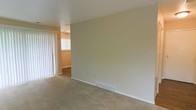 This single-story space presents a clean, straightforward layout with a focus on simplicity and functionality. The main living area is finished with wall-to-wall beige carpeting that lends a warm, understated feel underfoot. Neutral-toned walls in a soft cream palette keep the space bright and versatile, providing a blank canvas readily adaptable to various interior styles.

A floor-to-ceiling vertical blind treatment frames a sliding glass door or large window along one wall, allowing natural light to filter into the room while offering privacy control. The textured ceiling adds a subtle architectural detail consistent with traditional residential construction.

A floor-mounted HVAC vent along the baseboard indicates a central heating and cooling system, contributing to year-round comfort. A standard electrical outlet is conveniently positioned along the lower wall.

An open passageway connects the main living area to an adjoining room, which features a window that introduces additional natural light. The transition between spaces is defined by a subtle change in flooring, suggesting distinct functional zones within the single-floor layout. Overall, the space offers a practical, move-in-ready environment with a neutral and welcoming ambiance.