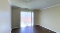 This single-story space features a clean, straightforward layout centered around a well-lit main living area. The room is finished with dark hardwood-style flooring that contrasts with the neutral, beige-toned walls, creating a warm and grounded atmosphere. Crown molding runs along the upper perimeter of the room, adding a subtle architectural detail that elevates the overall finish of the space.

The focal point of the living area is a large sliding glass door dressed with vertical blinds, which floods the interior with abundant natural light and provides direct access to an outdoor area. The bright exterior visible through the door suggests an open-air patio or balcony, offering a pleasant transition between indoor and outdoor living.

The ceiling features a textured popcorn finish, a common characteristic of this style of construction. White baseboards run along the base of the walls, providing a clean border between the flooring and walls. The overall ambiance of the space is bright, open, and functional, with minimal ornamentation that allows for flexible furnishing and personalization.