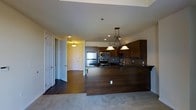 This single-floor residence showcases a thoughtfully designed open-concept layout that seamlessly connects the kitchen and living areas. The kitchen is a standout feature, boasting rich dark wood cabinetry that extends to the ceiling, complemented by dark granite countertops and a tile backsplash that adds texture and depth. Stainless steel appliances, including a refrigerator, microwave, and range, provide a sleek, modern touch. A generously sized kitchen island with a dark wood finish serves as both a functional workspace and a casual dining area, anchored by an elegant chandelier with warm pendant lighting that creates an inviting ambiance. Recessed lighting throughout ensures the space is well-illuminated. The flooring transitions from tile near the entryway to plush carpeting in the living area, offering both practicality and comfort. The entryway is framed by classic paneled doors and provides a clear sightline through the open living space. Overall, the residence combines warm, earthy tones with quality finishes to create a comfortable and functional living environment.