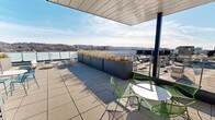 Located atop 2930 4th Ave S in Birmingham, Alabama, this rooftop terrace offers a sophisticated outdoor amenity space with sweeping panoramic views of the city skyline and the surrounding rolling hillside. The terrace is finished with large-format pavers and composite decking, providing a clean, modern aesthetic throughout.

The space is divided into distinct zones designed for both relaxation and social gathering. A partially covered section features a sleek paneled ceiling overhead, offering shade while maintaining an open, airy feel. This covered area is furnished with round bistro tables and colorful lattice-style chairs in green and teal, creating a vibrant, inviting atmosphere. A wall-mounted television is positioned in this covered zone, adding a functional entertainment element.

Large, rectangular planters filled with ornamental grasses and seasonal plantings are strategically placed across the terrace, adding greenery and serving as natural dividers between spaces. Open seating areas beyond the covered section take full advantage of the unobstructed sky and city views, with additional lounge seating and bistro arrangements dispersed throughout.

A sleek cable-rail perimeter railing with frosted glass panels ensures safety while preserving sightlines across Birmingham's urban landscape and horizon. The terrace receives abundant natural light throughout the day, making it an ideal venue for gatherings, events, or quiet outdoor relaxation.