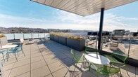 Located atop 2930 4th Ave S in Birmingham, Alabama, this rooftop terrace offers a sophisticated outdoor amenity space with sweeping panoramic views of the city skyline and the surrounding rolling hillside. The terrace is finished with large-format pavers and composite decking, providing a clean, modern aesthetic throughout.

The space is divided into distinct zones designed for both relaxation and social gathering. A partially covered section features a sleek paneled ceiling overhead, offering shade while maintaining an open, airy feel. This covered area is furnished with round bistro tables and colorful lattice-style chairs in green and teal, creating a vibrant, inviting atmosphere. A wall-mounted television is positioned in this covered zone, adding a functional entertainment element.

Large, rectangular planters filled with ornamental grasses and seasonal plantings are strategically placed across the terrace, adding greenery and serving as natural dividers between spaces. Open seating areas beyond the covered section take full advantage of the unobstructed sky and city views, with additional lounge seating and bistro arrangements dispersed throughout.

A sleek cable-rail perimeter railing with frosted glass panels ensures safety while preserving sightlines across Birmingham's urban landscape and horizon. The terrace receives abundant natural light throughout the day, making it an ideal venue for gatherings, events, or quiet outdoor relaxation.