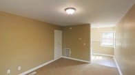This single-floor space is finished in warm tan/beige wall tones throughout, creating a cohesive and neutral aesthetic. The main room features wall-to-wall carpeting and is illuminated by a flush-mount ceiling light, giving the area a clean, simple feel. White painted baseboards provide a crisp contrast against the warm walls. A built-in white storage unit with upper cabinet doors and lower drawers is integrated into one wall, offering practical organization without the need for additional furniture. A floor-level HVAC vent is positioned nearby, indicating a built-in climate control system. An adjacent area, visible through an open passage, transitions to tile flooring and benefits from natural light streaming through a window, suggesting a secondary functional space such as a kitchen or utility area. A second flush-mount light fixture in this adjoining space ensures consistent lighting throughout. The overall design is straightforward and practical, emphasizing functionality with minimal ornamentation, making it a versatile space well-suited for a variety of uses.