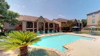 This single-story residential community presents an inviting and well-maintained exterior characterized by a warm beige and blue-gray color palette with arched architectural detailing. The covered front patio features rounded archways and louvered shutters, creating a shaded and welcoming entryway. Drought-tolerant landscaping with native grasses and yucca plants lines the walkway, adding a low-maintenance yet attractive curb appeal. A vintage Coca-Cola vending machine and casual outdoor dining furniture contribute a relaxed, community-friendly atmosphere to this area.

The centerpiece of the outdoor amenities is a generously sized swimming pool with striking turquoise water, framed by natural stone coping and a spacious sundeck. A row of bright teal lounge chairs lines the poolside, ideal for sunbathing, while additional shaded seating with umbrellas is available nearby. The multi-story residential buildings surrounding the pool feature pitched rooflines, open balconies with iron railings, and staircase access, all set against a backdrop of mature trees and abundant greenery. The overall ambiance is relaxed and resort-like, offering residents a comfortable outdoor retreat bathed in natural sunlight.