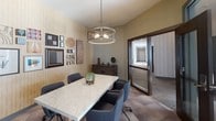 This thoughtfully designed single-floor space combines functionality with sophisticated interior styling, making it well-suited for meetings, collaboration, and community gathering. The centerpiece of the space is a private conference room enclosed by floor-to-ceiling glass walls and dark-framed sliding doors, offering both transparency and privacy. Inside, a marble-topped conference table is surrounded by upholstered charcoal chairs, anchored beneath a striking cylindrical drum pendant light with a metallic finish that adds a refined focal point overhead. One wall is finished in a textured, warm-toned wallcovering and serves as a curated gallery display, featuring an eclectic mix of framed artwork, mirrors, woven wall pieces, and decorative prints in varied sizes and styles, lending the room a creative and personalized character. A dark wood credenza along the adjacent wall provides additional storage and display surface. The flooring throughout features a geometric tile pattern in muted tones, adding visual interest underfoot. Beyond the conference room, a corridor leads to a bright and airy communal lounge filled with natural light streaming through large windows that frame outdoor views. This lounge area features hardwood flooring, dining-style seating, pendant lighting, and an open, welcoming layout ideal for casual gatherings or shared use.