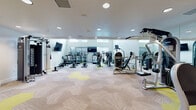 This fitness center is a comprehensively outfitted facility designed to support a wide range of workout routines. The space is organized across a single level and divided into distinct functional zones, each tailored to specific exercise needs.

The cardio area is generously sized and houses an array of machines, including treadmills, stationary bikes, ellipticals, stair climbers, and rowing machines. Wall-mounted flat-screen televisions are positioned throughout for entertainment during workouts. Large windows along one wall allow abundant natural light to fill the space, while clerestory windows add additional brightness near the ceiling.

The strength training section features a cable machine system, weight benches, and a full dumbbell rack with accessory equipment such as medicine balls and stability balls. Floor-to-ceiling mirrors line the walls, enhancing the sense of space and allowing users to monitor their form.

A standout feature is the enclosed children's play area, accessible via glass-panel doors. This compact, thoughtfully designed room includes child-sized furniture, colorful play mats, and a mounted television, making it convenient for caregivers.

Throughout the facility, the flooring consists of a distinctive patterned carpet in neutral tones accented with yellow-green geometric shapes, contributing to the modern, clean aesthetic of the overall space.