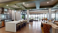 This thoughtfully designed single-floor amenity space makes a striking first impression through a bold entrance foyer clad in dark, textured wall paneling, flanked by sleek black planters with lush greenery and large-format tile flooring. Double glass doors open into a spacious, open-concept interior defined by coffered ceilings with rich green and charcoal accents, recessed lighting, and warm hardwood-style flooring throughout.

The gourmet kitchen is a standout feature, equipped with a professional-grade stainless steel range, a dramatic stone tile backsplash, a powerful overhead hood, and a generous island with a white quartz countertop, undermount sink, and bar seating. Pendant lighting adds warmth and visual interest above the counter.

The main lounge area is anchored by a stone-clad fireplace wall with a mounted flat-screen television, surrounded by plush neutral sofas, leather accent chairs, and vibrant mustard-toned throw pillows. A separate social seating zone features an expansive sectional sofa and additional lounge chairs arranged around a sculptural wood coffee table on a bold area rug.

Floor-to-ceiling windows and glass doors flood the interior with natural light and provide seamless access to an outdoor terrace, creating an inviting indoor-outdoor connection throughout the space.