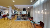 This thoughtfully designed single-floor community space offers a rich variety of amenity areas that cater to both relaxation and socializing. The entrance corridor features warm hardwood-style flooring, deep slate-blue accent walls, and recessed lighting that creates a polished, welcoming atmosphere leading residents into the main common areas.

The expansive central gathering space is anchored by a large kitchen with a generous island, white quartz countertops, light wood cabinetry, a black appliance suite, and a mosaic tile backsplash. Pendant lighting over the island adds a refined touch, while the open layout connects seamlessly to a game and recreation area complete with a billiard table and a tiled fireplace.

The lounge area features comfortable leather and upholstered seating, striking large-drum pendant light fixtures, an inset television, and a decorative etched-glass water feature wall that adds a distinctive architectural element. Natural light pours in through floor-to-ceiling windows draped in sheer curtains, creating bright, airy dining nooks with intimate table settings and direct views to the outdoors.

Throughout the space, warm hardwood flooring, varied ceiling heights with contrasting color treatments, and carefully curated furnishings combine to create an inviting, community-focused environment that balances functionality with contemporary design.