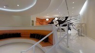 This visually captivating space makes an immediate impression through its bold, avant-garde design language. The interior is dominated by smooth, flowing curved walls and ceilings rendered in crisp white, creating a futuristic aesthetic that feels both dynamic and cohesive. A sweeping circular ceiling recess with integrated lighting adds depth and drama overhead, while recessed spotlights throughout ensure the space is well-illuminated.

A standout feature is the large-scale black and white mural depicting a dramatic, expressive face with flowing hair set against a vivid orange accent wall — a striking artistic centerpiece that anchors the space visually. This bold color contrast between the white structural elements and the warm orange tones infuses the interior with energy and personality.

A curved, built-in orange sofa with dark accent cushions echoes the organic forms of the architecture, creating a comfortable and stylish seating area that integrates seamlessly into the flowing design. A sleek white staircase with a clean railing design adds a sculptural element while connecting different levels of the space.

Pendant light fixtures hang at considered intervals, contributing to the warm, curated ambiance. Overall, the space exudes a contemporary, design-forward character suited to a boutique, hospitality, or high-end commercial environment.
