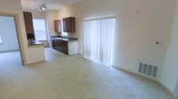 This single-floor space showcases an open-concept design that seamlessly connects the kitchen and living areas into one cohesive, functional layout. The living area is finished with neutral-toned carpeting that adds warmth underfoot, while the walls are painted in a soft, light beige that enhances the sense of openness and brightness throughout.

The kitchen is positioned along one wall and features dark-stained wood cabinetry that provides a striking contrast against the lighter surroundings. A double-basin sink sits beneath the cabinetry, complemented by dark countertops that offer a practical workspace. A track lighting fixture above the kitchen area ensures ample task lighting, while a window to the left of the kitchen invites additional natural light into the space.

Dominating the far wall of the living area is a large sliding glass door dressed with vertical blinds, which serves as the primary source of natural light and connects the interior to an outdoor area. The transition between the carpeted living space and the tiled kitchen area is clean and well-defined. Overall, the space offers a simple, functional layout with a neutral, understated aesthetic well-suited for everyday living.