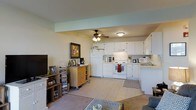 This single-floor space showcases an open-concept design that integrates the living area and kitchen into one cohesive, functional layout. The kitchen is well-appointed with white cabinetry extending to the upper walls, providing generous storage throughout. A white electric range and oven serve as the central cooking appliance, complemented by a refrigerator and a microwave positioned on the countertop. The counters offer a practical workspace, and a decorative rug anchors the kitchen area. Light tile flooring runs throughout the kitchen and transitions into carpet within the living space, subtly defining each zone. A ceiling fan with a light fixture is centrally mounted, ensuring air circulation and supplemental lighting across the shared space. The living area accommodates a television, a media storage unit, and a rolling wooden cart that adds a casual, flexible storage solution. A framed corkboard on the wall adds a personal touch. The overall ambiance is casual and functional, with a neutral color palette of whites and soft tones creating a clean, approachable atmosphere throughout.