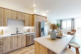 This thoughtfully designed one-bedroom, one-bathroom residence offers a comfortable and cohesive living experience across a single floor. The open-concept kitchen and living area forms the heart of the home, where warm wood-tone cabinetry with matte black hardware, white quartz countertops, and a full suite of stainless steel appliances create a functional yet stylish kitchen. A central island with bar seating provides additional prep space and casual dining. Light wood-style flooring flows throughout, unifying the space with a clean, contemporary aesthetic.

The living area is bright and inviting, with large windows and a sliding glass door that welcome natural light and provide outdoor views. The bedroom offers a comfortable retreat, highlighted by a statement accent wall with textured wallcovering, a large window with neighborhood views, and light wood flooring. A dedicated closet provides practical storage.

The bathroom is well-appointed with a wood-tone vanity, white quartz countertop, large-format tile flooring, and a spacious walk-in glass-enclosed shower featuring marble-style tile surround and matte black fixtures. A hallway connects the rooms, completing this efficient and modern layout.