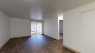 This single-floor unit offers a straightforward, functional layout that makes efficient use of its space. The living area serves as the focal point of the unit, featuring rich, dark hardwood-style flooring that runs throughout. Crisp white walls and a smooth ceiling create a clean, neutral backdrop, while a sliding glass door floods the room with natural light and provides direct access to an outdoor patio area enclosed by a privacy fence. A wall-mounted heater unit is also present in the living space, adding a practical element to the room.

An open passage connects the living area to the kitchen, which is equipped with white appliances including a coil-burner electric range with overhead ventilation, a full-size refrigerator, and a double-basin sink. Wood-trimmed cabinetry in a warm brown tone lines the upper and lower walls, complemented by light-toned laminate countertops that provide ample prep space.

The bathroom continues the cohesive hardwood-style flooring found throughout the unit and includes a wood-trimmed vanity with a light countertop, a wall-mounted towel bar, and standard fixtures. The overall ambiance of the unit is clean and understated, offering a move-in-ready space with a neutral palette suitable for a variety of furnishing styles.