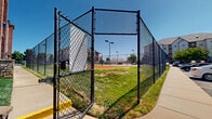 Located at 2020 E Kerr St in Springfield, Missouri, this outdoor recreational complex offers a well-rounded range of amenities designed for active use and community engagement. The centerpiece of the space is a full-sized tennis court with a regulation net, a smooth hard surface, and clearly marked lines, complemented by dedicated lighting poles that allow for use during evening hours. Adjacent to the tennis court is a multi-use basketball court, also outfitted with a backboard and hoop, regulation court markings, and a textured, interlocking tile surface built for durability and performance. Both courts are fully enclosed by black chain-link fencing with gated access points, providing a secure and defined playing environment. A children's playground area sits nearby, featuring a multi-component play structure with slides and climbing elements, set within a safety-surfaced enclosure bounded by its own chain-link perimeter. Open grassy areas and paved walkways connect the various recreational zones, creating a cohesive outdoor layout. The surrounding multi-story residential buildings are visible from throughout the complex, and the facility benefits from generous natural light and open sky views. Ample on-site parking is accessible along the perimeter, making the complex convenient for regular use.