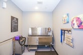 This single-floor pet spa is purpose-built to provide a thorough and comfortable grooming experience for dogs. The space is finished with neutral gray walls and durable, easy-to-clean flooring, creating a hygienic and welcoming environment. At the heart of the room is a large, deep stainless steel bathing tub equipped with a mounted sprayer and retractable door for easy pet access. Adjacent to the tub is a professional-grade forced-air pet blower, ideal for efficiently removing excess water and loose fur from a pet's coat following a bath. A grooming table with a non-slip rubber mat surface and an attached grooming arm provides a stable and secure station for post-bath care. A leash rack mounted on the wall offers organized storage for leads during visits, accompanied by cheerful canine-themed artwork and a welcoming decorative sign that adds a warm, pet-friendly character to the space. A clearly posted "Pet Spa Rules" sign ensures all guests understand proper equipment usage and etiquette. The room connects through a white paneled door to an adjacent hallway with wood-look flooring, allowing for a smooth flow throughout the facility. Overall, the space balances functional grooming infrastructure with an inviting, dog-loving aesthetic.