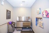 This single-floor pet spa is purpose-built to provide a thorough and comfortable grooming experience for dogs. The space is finished with neutral gray walls and durable, easy-to-clean flooring, creating a hygienic and welcoming environment. At the heart of the room is a large, deep stainless steel bathing tub equipped with a mounted sprayer and retractable door for easy pet access. Adjacent to the tub is a professional-grade forced-air pet blower, ideal for efficiently removing excess water and loose fur from a pet's coat following a bath. A grooming table with a non-slip rubber mat surface and an attached grooming arm provides a stable and secure station for post-bath care. A leash rack mounted on the wall offers organized storage for leads during visits, accompanied by cheerful canine-themed artwork and a welcoming decorative sign that adds a warm, pet-friendly character to the space. A clearly posted "Pet Spa Rules" sign ensures all guests understand proper equipment usage and etiquette. The room connects through a white paneled door to an adjacent hallway with wood-look flooring, allowing for a smooth flow throughout the facility. Overall, the space balances functional grooming infrastructure with an inviting, dog-loving aesthetic.