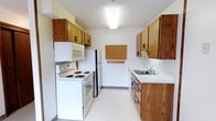 This single-floor space is designed with functionality and practicality in mind. The kitchen is laid out in a classic galley configuration, with warm oak wood cabinetry lining both sides to maximize storage. Upper and lower cabinets provide ample space for pantry and kitchen organization, while white laminate countertops offer a clean, bright working surface. The kitchen is fully equipped with a white electric coil-top range, an over-the-range microwave, and a full-size refrigerator. A single-basin stainless steel sink with a standard faucet is positioned beneath additional cabinetry on the opposite wall, complemented by a subtle tile backsplash. A cork bulletin board mounted on the wall between the counters adds a practical touch for notes and reminders, alongside a small wall-mounted key and accessory hook. The flooring throughout is a light-toned vinyl, easy to maintain and consistent with the space's straightforward aesthetic. Overhead, a flush-mount fluorescent light fixture provides bright, even illumination across the kitchen. The overall ambiance is clean and utilitarian, making efficient use of the available space.