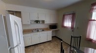 This single-story space includes a kitchen that serves as a central functional area. The kitchen is outfitted with white upper and lower cabinetry, providing ample storage throughout. A double-basin sink sits beneath the upper cabinets, accompanied by a light-toned laminate countertop that extends along the wall. A dedicated built-in desk or utility counter area is positioned adjacent to the main countertop, offering additional workspace. A microwave is placed on the countertop extension, and a small decorative plant adds a subtle touch of greenery to the upper shelving. The flooring consists of tile in a warm earth tone, contributing to a clean and practical aesthetic. Natural light enters the kitchen through a window dressed with pink/mauve curtains, adding a soft accent to the otherwise neutral white and beige palette. The walls are painted in a light neutral tone, maintaining a simple and straightforward interior style. An entry door is visible near the window, suggesting this kitchen area is positioned close to an exterior access point of the space.