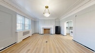 Located at 600 James St in Syracuse, NY, Unit 101 is a thoughtfully laid-out single-floor residence that blends classic architectural character with practical functionality. The living room serves as the centerpiece of the unit, showcasing ornate crown molding, elegant wainscoting panels, and a decorative white fireplace with detailed tile surround. Light oak hardwood flooring flows throughout, complemented by soft blue-gray walls and a brass pendant light fixture that adds a warm, vintage touch. Dual windows fitted with decorative radiator covers allow natural light to fill the room, creating an airy and inviting atmosphere. The entry area continues the refined detailing with intricate crown molding and a solid wood front door. The kitchen is efficiently designed, featuring white cabinetry, a stainless steel sink with marble-style countertops, and an electric range — providing all essential cooking amenities. The bathroom is clean and functional, with white tile walls, a walk-in shower stall, and light wood flooring. An in-unit laundry area adds everyday convenience. Overall, the space offers a compelling mix of historic architectural charm and updated practical features in the heart of Syracuse.