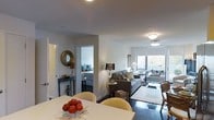 Located at 10 DeKalb Ave in White Plains, New York, this single-floor residence offers a well-designed, open-concept layout that seamlessly connects the main living areas. The dining area anchors one end of the space, featuring a clean white table surrounded by curved cream-toned chairs, creating an inviting setting for meals and gatherings. A decorative bowl of fruit adds a warm, welcoming touch to the tablescape.

The living room flows naturally from the dining area, centered around a plush gray sofa and accented by warm table lamps and a patterned area rug over rich dark hardwood flooring. Floor-to-ceiling windows flood the space with abundant natural light and provide views of the outdoors, enhancing the open and airy atmosphere.

The bedroom, visible through a connecting doorway, is appointed with a neatly made bed and coordinating blue accent pillows, while a decorative gold mirror and a classic console table in the entryway area contribute to the space's refined aesthetic.

The interiors throughout are finished in crisp white walls with smooth ceilings, complemented by flush and pendant lighting fixtures. The overall ambiance is modern yet comfortable, striking a balance between elegant design and everyday livability.