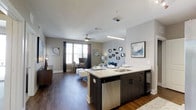 This single-floor residence showcases a thoughtfully designed open-concept layout that seamlessly connects the kitchen and living areas. The kitchen is anchored by a large island with a white quartz-style countertop, an undermount double sink, dark cabinetry, and stainless steel appliances, including a dishwasher and a beverage refrigerator. Pendant lighting above the island adds a contemporary touch, while the open sightlines create a welcoming and functional space for entertaining.

The living area extends naturally from the kitchen, featuring warm hardwood-style flooring that runs throughout the space, contributing to a cohesive and polished aesthetic. A ceiling fan with a light fixture provides both comfort and illumination, while large windows dressed with dark curtains allow natural light to fill the room. The neutral gray wall palette enhances the bright, airy ambiance. Decorative wall accents, including circular mirrors and framed artwork, add character to the space. Overall, the layout balances style and practicality, offering a comfortable and modern living environment.