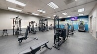 This 910 square foot exercise room, located at 3549 Iowa Ave, Riverside, CA, is a comprehensive fitness facility designed to cater to a wide range of workout preferences. The space features a well-planned layout with distinct zones for various exercise types. The room is equipped with state-of-the-art fitness machines, including treadmills, ellipticals, and stationary bikes for cardiovascular training. A dedicated strength training area includes weight machines, free weights, and a power rack for resistance exercises. The facility also incorporates functional training equipment and open floor space for stretching or bodyweight exercises. Motivational wall art with fitness-related words adds an inspiring atmosphere to the environment. The room's design includes fluorescent lighting, a dropped ceiling, and gray speckled flooring, creating a clean and modern aesthetic. Large windows allow natural light to enter, enhancing the overall ambiance. French doors and additional entryways provide easy access and contribute to the open feel of the space. This well-appointed exercise room offers a complete fitness solution in a single, efficiently designed area.