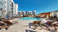 Located at 1880 Maple Dr NW in Kennesaw, Georgia, this outdoor amenity space is designed around a generously sized resort-style swimming pool that serves as the centerpiece of the area. The crystal-clear pool features an eye-catching water fountain feature and is bordered by a wide, stone-paved deck finished in natural-toned pavers that add warmth and texture to the surroundings.

Ample lounge seating lines the pool deck, offering residents plenty of space to relax and enjoy the open sky. Elegant wood-beam pergolas are positioned at key points around the pool, providing shaded retreats and defining the architectural character of the outdoor space. A dedicated outdoor kitchen and grilling station equipped with multiple built-in grills is situated along the perimeter, making it ideal for social gatherings.

The pool area is enclosed by a decorative iron fence, ensuring both security and an unobstructed view of the surrounding landscape, which includes mature trees and neighboring residential buildings. The multi-story residential buildings flanking the space feature modern facades with mixed materials including board-and-batten siding and large windows with private balconies. The overall ambiance is one of a well-appointed, community-focused outdoor retreat that balances functionality with upscale design.