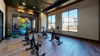 Located on Poinsett Way in Pooler, Georgia, this single-floor fitness studio is purpose-built for group cycling and functional training. The space is thoughtfully designed with a refined yet energetic aesthetic, combining wood-look plank flooring, wood-paneled accent walls, and a deep green tray ceiling accented by recessed lighting, wall sconces, and a ceiling fan for enhanced air circulation during workouts.

A row of stationary cycling bikes occupies the central floor space, providing ample room for group spin sessions. Along one wall, a large-scale abstract watercolor mural serves as a striking focal point, adding an artistic dimension to the workout environment. Full-length mirrors and glass-paneled interior doors expand the sense of space and allow visual connectivity with adjacent areas.

Natural light floods the studio through oversized black-framed windows, offering views of the surrounding community and landscape. A dedicated storage rack holds an assortment of colorful exercise balls and resistance equipment, supporting a range of fitness activities beyond cycling. The overall ambiance balances functionality with contemporary design, creating a motivating and visually engaging environment for residents and users.
