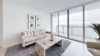 Unit 49E is a meticulously designed high-rise residence that blends modern minimalism with stunning panoramic water views. The open-concept living area is anchored by floor-to-ceiling sliding glass doors that lead to a private balcony, flooding the space with natural light and framing breathtaking cityscape and waterfront vistas. Light-toned wood flooring runs throughout the main living areas, complementing the crisp white walls and creating a clean, airy aesthetic.

The kitchen is outfitted with high-gloss white cabinetry, dark stone countertops, stainless steel Bosch appliances, and a central island — combining functionality with a refined, contemporary look. Pendant lighting above the island adds a polished finishing touch.

The bedroom is generously proportioned, featuring the same light wood flooring and direct access to the balcony through additional floor-to-ceiling glass doors, continuing the theme of seamless indoor-outdoor living.

The bathroom showcases marble-look tile flooring, a sleek white floating vanity with modern hardware, a large mirror, and a glass-enclosed shower, delivering a spa-like experience. Throughout the residence, the consistent use of white tones, clean lines, and quality materials creates a cohesive and sophisticated atmosphere.
