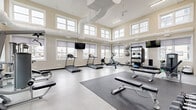 Located at 5000 Green Jewel Blvd in Regina, Saskatchewan, this fitness facility is a thoughtfully designed, single-floor gym that offers a spacious and welcoming environment for a full range of workouts. The room features impressively high ceilings with a two-tiered window arrangement, allowing natural light to flood in from multiple directions and creating an open, airy atmosphere. Warm neutral-toned walls complement the clean, light-coloured flooring, with rubberized mat zones strategically placed for strength and stretching activities. Ventilation ductwork runs visibly along the ceiling, supported by a ceiling fan that ensures comfortable airflow throughout. The cardio section is well-stocked with multiple treadmills, an elliptical trainer, and a rowing machine, all positioned along the windowed walls to take advantage of the natural light and outdoor views. A dedicated strength training area includes adjustable benches, cable machines, a dumbbell rack, and a variety of free weights and medicine balls. Large wall-mounted mirrors on one side of the room enhance the sense of space and allow users to monitor their form. Multiple flat-screen televisions are mounted at convenient viewing angles throughout the facility. The gym is clean, bright, and efficiently organized to support diverse fitness routines.