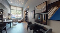 This thoughtfully designed single-floor space is organized into several distinct yet cohesive zones, united by light hardwood flooring, high ceilings, and a polished contemporary aesthetic. The entry area makes a strong first impression with large floor-to-ceiling glass doors, frosted glazing panels, and a dramatic textured accent wall featuring a bold geometric relief pattern. Gold-framed open shelving styled with curated books and decorative objects adds warmth to the arrival experience.

The library and lounge area is bathed in natural light from oversized windows overlooking mature trees outdoors. Built-in bookshelves line the walls, while a sculptural ceiling installation of clustered recessed spotlights adds visual interest overhead. Dark tables with upholstered seating offer workspace, complemented by a cozy reading nook with an accent chair and ottoman. Large-format architectural photography adorns the walls.

The game and entertainment lounge takes on a dramatically different character, with deep charcoal walls, bold oversized drum pendants, and a tufted leather sofa accented with chartreuse pillows. A grid of black-and-white music-themed artwork anchors the space, while a wall-mounted television and a striking red billiard table — lit by woven pendant fixtures — complete the energetic, social atmosphere.