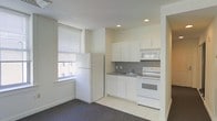 This single-floor unit features an open, functional layout with a kitchen area that transitions seamlessly from the main living space. The kitchen is outfitted with white cabinetry, offering ample storage through both upper and lower cabinets. A gray tile backsplash adds a subtle contrast against the white surfaces, while the countertop provides practical workspace. Essential appliances are included, featuring a white refrigerator, electric range with overhead microwave, and a single-basin sink. Recessed lighting in the kitchen area ensures well-lit food preparation. The living area is finished with dark carpeting, creating a visual distinction from the light tile flooring of the kitchen. A window fitted with horizontal blinds allows natural light to fill the space while maintaining privacy. The neutral, light-toned walls throughout contribute to a clean, understated ambiance. The layout is straightforward and efficient, making practical use of the available space across the single floor.