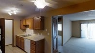 This single-story space presents a practical and cohesive layout designed for everyday living. The kitchen is equipped with warm-toned wood cabinetry, offering ample upper and lower storage. Light countertops provide a clean contrast to the rich cabinetry, and a white dishwasher and sink are neatly integrated beneath the counter. A ceiling fan with a light fixture overhead keeps the space well-ventilated and illuminated. The textured ceiling adds subtle architectural character throughout. An open passageway connects the kitchen directly to the adjacent carpeted living area, creating a natural flow between spaces. The living area benefits from natural light streaming through a window, brightening the neutral-toned walls. A bifold closet door is visible near the entryway to the living space, offering convenient storage. The consistent use of neutral wall colors and warm wood tones throughout creates a unified, comfortable ambiance suited for practical, day-to-day living.