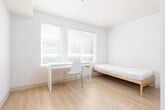This single-floor studio unit offers a thoughtfully organized layout that maximizes functionality within a compact footprint. The interior features a clean, minimalist aesthetic throughout, with white walls, smooth ceilings, and warm light wood laminate flooring that runs continuously across the main living areas, creating a cohesive and open feel.

The sleeping area accommodates a full-size bed frame and is positioned alongside a window fitted with horizontal blinds, allowing for natural light and outdoor views while maintaining privacy. Adjacent to the sleeping area is a dedicated workspace, anchored by a built-in white desk stretching beneath two large windows. This corner is particularly well-lit with natural light streaming in from multiple angles, making it well-suited for study or remote work.

The kitchenette is neatly integrated near the entry and includes a countertop with a stainless steel sink, open wall-mounted shelving for storage, and space for a microwave. The entry door features a secure electronic lock, reinforcing the unit's practicality.

The bathroom is tiled in neutral beige tones and is equipped with a toilet, wall-mounted sink, and a grab bar, indicating accessibility-conscious design. An electrical panel is conveniently located just outside the bathroom. Overall, the unit presents a clean, move-in-ready space suited for a single occupant seeking a straightforward, low-maintenance living environment.
