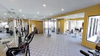 This fitness center offers a thoughtfully designed workout environment spread across a single floor. The space is defined by warm yellow walls, recessed lighting, and a checkered gray-and-white tile floor that lends a clean, functional aesthetic throughout. Large windows along one wall allow natural light to filter in, creating a bright and energizing atmosphere.

Floor-to-ceiling mirrors are strategically positioned to visually expand the room and allow users to monitor their form during exercise. The facility is equipped with a variety of strength training machines, including cable pulley systems and resistance equipment, as well as cardio machines such as an elliptical trainer. The equipment is arranged to allow comfortable circulation and use of multiple stations simultaneously.

Recessed ceiling lights provide consistent, even illumination throughout the space, complementing the natural light from the windows. A standard entry door is visible toward one end of the room, and a trash receptacle is conveniently placed nearby. Overall, the fitness center presents a practical and well-maintained environment suited for a range of workout routines.