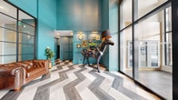 This distinctive building lobby makes an immediate impression with its soaring double-height ceilings and vibrant teal walls that set a bold, artistic tone throughout the space. The floor is finished in a dramatic large-format herringbone pattern, combining grey and neutral tones to create a visually dynamic foundation that anchors the entire area.

The main lounge area features a pair of richly upholstered brown Chesterfield sofas and a tufted leather armchair arranged around a gold-accented side table, offering a comfortable and stylish seating arrangement for residents and guests. Tall floor-to-ceiling glazed walls and sliding glass entry doors flood the lobby with abundant natural light, while sheer and draping curtains add softness to the expansive windows.

A standout decorative element is the life-sized sculptural black horse fitted with a saddle and functioning as a lamp with a bronze-lined shade — an unmistakable artistic centerpiece. Curated framed artwork displayed on paneled teal walls and potted tropical plants add further character and warmth to the space.

Adjacent to the main lobby, a separate mail room area is finished with warm wood cabinetry, a clean white countertop, rows of individual mailboxes, and pendant lighting — providing a functional yet refined utility zone. The overall ambiance of the lobby is one of contemporary sophistication blended with eclectic, gallery-inspired design.