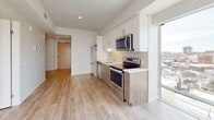 Located on the 10th floor at 63 Scott St in Kitchener, Ontario, this thoughtfully designed unit offers a clean, contemporary aesthetic across its single-floor layout. Light-toned hardwood-style flooring runs throughout the space, creating a sense of continuity and warmth from room to room.

The kitchen is a standout feature, outfitted with a full suite of stainless steel appliances including a bottom-freezer refrigerator, electric range with microwave, and dishwasher. Two-tone cabinetry pairs white upper cabinets with warm wood-finish lower cabinets, complemented by a white subway tile backsplash and light quartz countertops. A stainless steel sink sits beneath a window, and an adjacent floor-to-ceiling window fills the space with natural light and urban views.

The bedroom is clean and minimalist, featuring crisp white walls and a large window with a glass door that opens to an outdoor-facing view of the surrounding cityscape, allowing abundant natural light to pour in.

The bathroom is finished with large-format neutral tile on both the floors and tub surround, housing a soaker tub with a curtain rod, a toilet, and a grab bar — combining functionality with a calm, spa-like atmosphere.

Two hallways connect the rooms efficiently, and the overall layout makes excellent use of the available footprint, delivering a modern, move-in-ready living space in the heart of Kitchener.