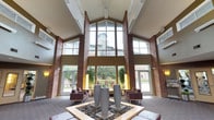 This single-floor lobby space makes a striking first impression with its grand atrium entrance, characterized by soaring ceilings, floor-to-ceiling windows framed by exposed brick columns, and an abundance of natural light. A central decorative installation featuring river rock and tall cylindrical planters anchors the seating area, which is furnished with deep plum upholstered chairs arranged to encourage conversation. The expansive glazing offers clear views of the surrounding outdoor landscape, further enhancing the open, airy atmosphere.

A secondary entry vestibule connects seamlessly to the main lobby, featuring rich stone tile flooring with a curved design motif, warm amber and dark chocolate wall tones, and a pendant light fixture that adds a refined touch. This area provides a welcoming seating nook with contemporary furnishings positioned near the windows.

The lobby corridor winds gracefully with curved walls finished in dark wood paneling and neutral tones, with recessed lighting illuminating the path. Artwork accents the hallway, adding a cultured ambiance. Adjacent to the elevator bank, a dedicated mailroom offers a wall of individual resident mailboxes. The elevator lobby showcases polished stainless steel doors set against dark stone tile surrounds, complemented by a bold overhead pendant light, completing the building's sophisticated and cohesive design aesthetic.