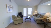 This single-floor space centers around a welcoming living room designed for both comfort and functionality. The room features neutral-toned walls and wall-to-wall carpeting that creates a warm, cohesive atmosphere throughout. Elevated ceilings add a sense of openness, while a centrally placed glass-panel door and an adjacent window allow natural light to fill the space and provide a view of the outdoors.

The living area is furnished with two sage-green upholstered sofas — a full sofa and a matching armchair — arranged around a dark wood coffee table, creating an inviting seating arrangement. A flat-screen television is positioned on a low media console along one wall, offering a practical entertainment setup. A framed artwork piece adorns the wall, adding a subtle decorative touch. A table lamp near the sofa provides warm ambient lighting for evening use.

The layout is straightforward and efficient, with clear sightlines across the room. The combination of soft carpeting, neutral walls, and ample natural light gives the space a clean, livable quality suited for everyday use.