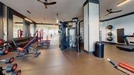 Located at 55 N Park Ave in Tucson, Arizona, this fitness center offers a generously sized, single-floor exercise facility designed to accommodate a comprehensive range of workout styles. The space is organized into distinct zones, providing a well-structured environment for both strength and cardiovascular training.

The strength training area is outfitted with a multi-functional cable machine system, a full rack of dumbbells along a mirrored wall, adjustable benches, and a variety of selectorized weight machines with red upholstered seating. A dedicated functional fitness zone features wall-mounted medicine balls, TRX suspension trainers, and resistance bands, offering ample space for dynamic, open-floor exercises.

The cardio section benefits from dramatic floor-to-ceiling windows that flood the area with natural light and offer views of the surrounding Tucson landscape. It houses a row of elliptical trainers, stair climbers, and treadmills. Ceiling fans and pendant lighting throughout maintain comfort and a modern aesthetic.

The facility features durable rubber flooring throughout, high ceilings that create an open and airy atmosphere, and motivational signage on the walls. Large glass partitions between zones maintain visual connectivity while defining each workout area. The overall design balances functionality with a clean, modern aesthetic suited for serious fitness use.