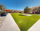 This unique space seamlessly blends indoor and outdoor elements. The interior features a single room of approximately 160 square feet, with cream-colored walls and a smooth, light-colored floor. A standout feature is the decorative window with a geometric pattern, framed by white trim, which adds character and allows natural light to filter in. The room is equipped with recessed lighting and appears to have climate control capabilities.

The space opens directly onto an expansive, well-maintained lawn area. This green space is a significant feature, providing a stark contrast to the indoor area. The lawn is surrounded by Mediterranean-style buildings with terracotta roofs, creating a picturesque, resort-like atmosphere. Mature trees line the perimeter, offering shade and natural beauty.

The transition between indoor and outdoor is facilitated by a wide opening, possibly sliding doors, allowing for an easy flow between the two areas. This design maximizes the usable space and creates a seamless indoor-outdoor living experience. The overall effect is one of openness and connection to nature, despite the compact interior dimensions.